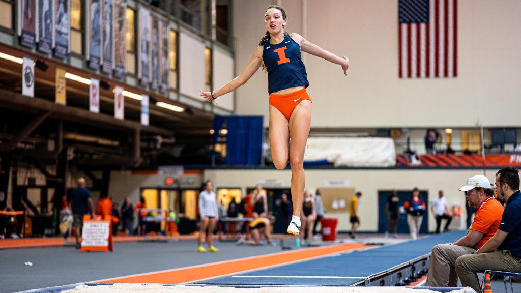 Darja Sopova is triple jumping down a long dark blue floor. She is airborne in a blue and orange uniform. 