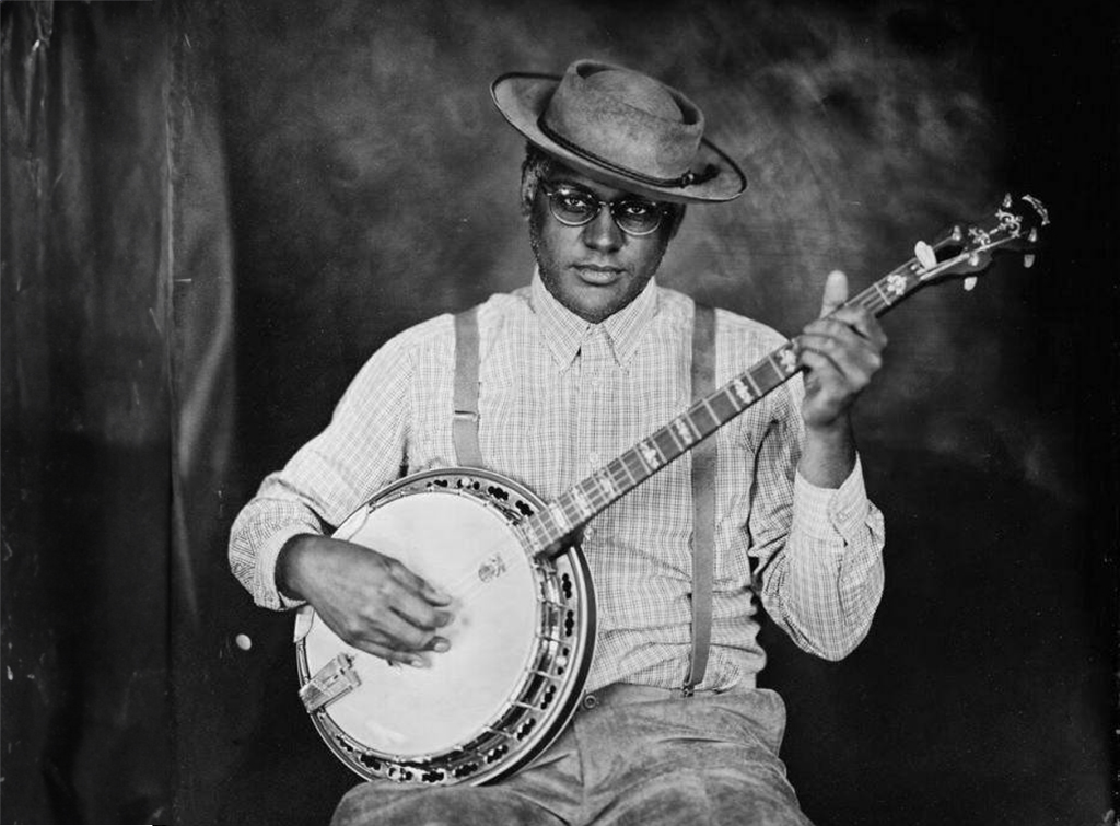 A person is seated, wearing a light-colored, long-sleeved, button-up shirt and dark pants. Suspenders stretch over the shoulders. A wide-brimmed hat rests on the head. The individual is playing a banjo, their fingers poised mid-strum. The image is in black and white, and the person sits against a dark backdrop. The person holds a banjo, their hands positioned as if playing it. The background is dark and nondescript. 