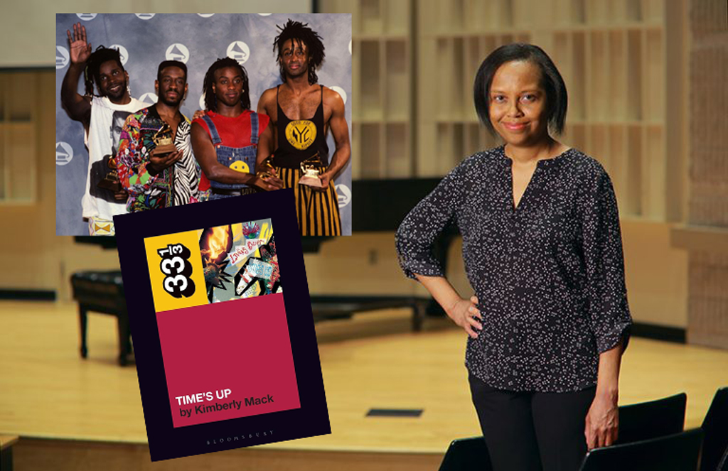In the image, a woman stands confidently in a room that appears to be a lecture hall or auditorium. She is dressed in a dark, floral-patterned blouse with a rounded neckline and three-quarter length sleeves. In the background, there's a collage of images superimposed over the scene: on the left, a group of four men is holding Grammy awards, celebrating their victory. Below them is a cover of a book or an album titled "TIME'S UP" by Kimberly Mack, featuring a colorful, abstract design. The atmosphere conveys a sense of academic and musical achievement.