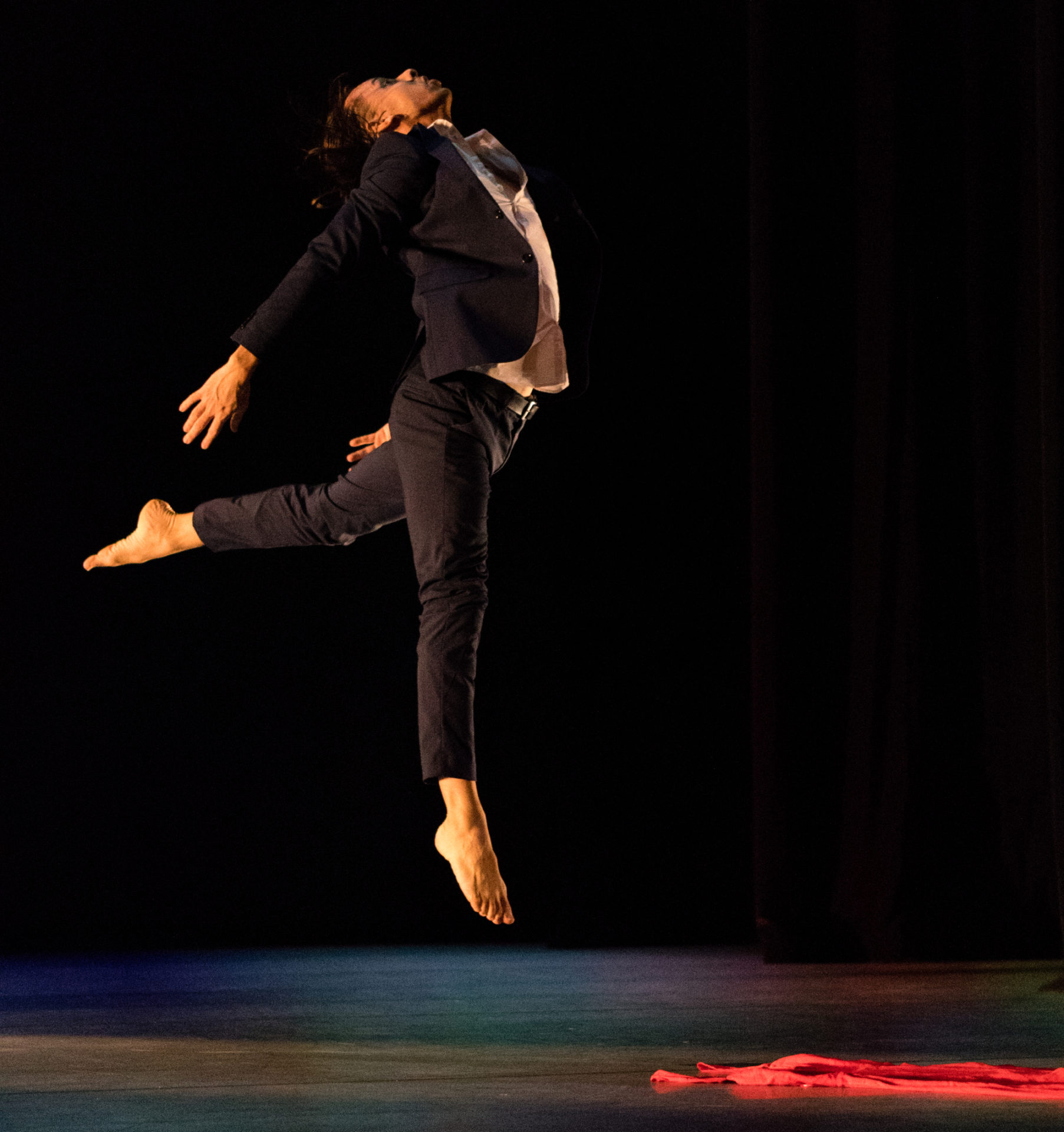 A Black man dancer is captured mid-jump. His left leg is kicked back and his right leg is straight, but off the ground. His back is arched, arms are back, and head is tilted toward the ceiling. There is red fabric on the stage.