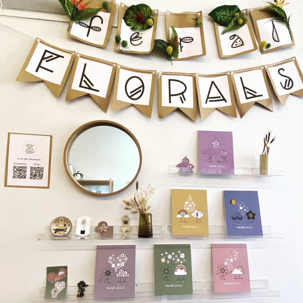 A white wall with a feral floral banner stretched across and shelves with colorful cards and stickers. There is a small circular mirror on the left side.