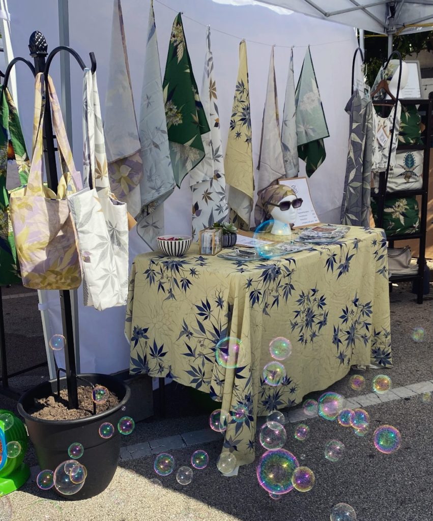 A market set up of feral florals merchandise. There are tablecloths, purses, and bandanas with bubbles in the bottom of the image. 