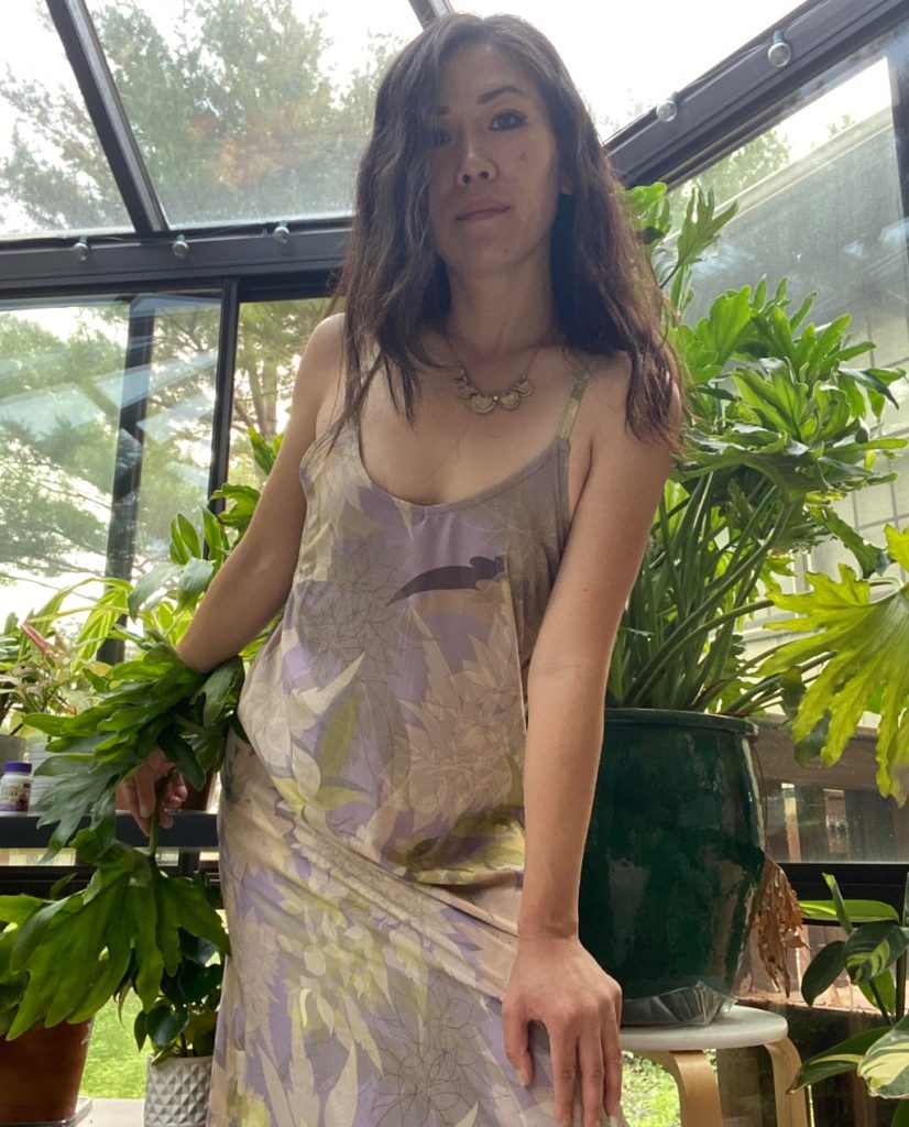 Janice Weldon stands in front of a large green bush in a greenhouse, slightly above the camera looking down. She is wearing a mauve dress with a cannabis leaf. She has dark brown hair iand a her hand on her knee. 
