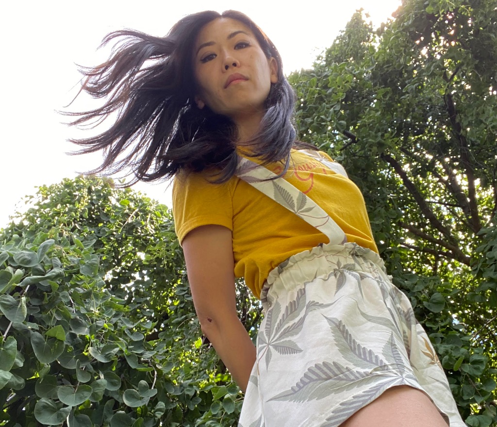 Janice Weldon stands in front of a large green bush, slightly above the camera looking down. She is wearing a yellow t-shirt and a leaf patterned romper. Her dark brown hair is blowing slightly in the wind. 
