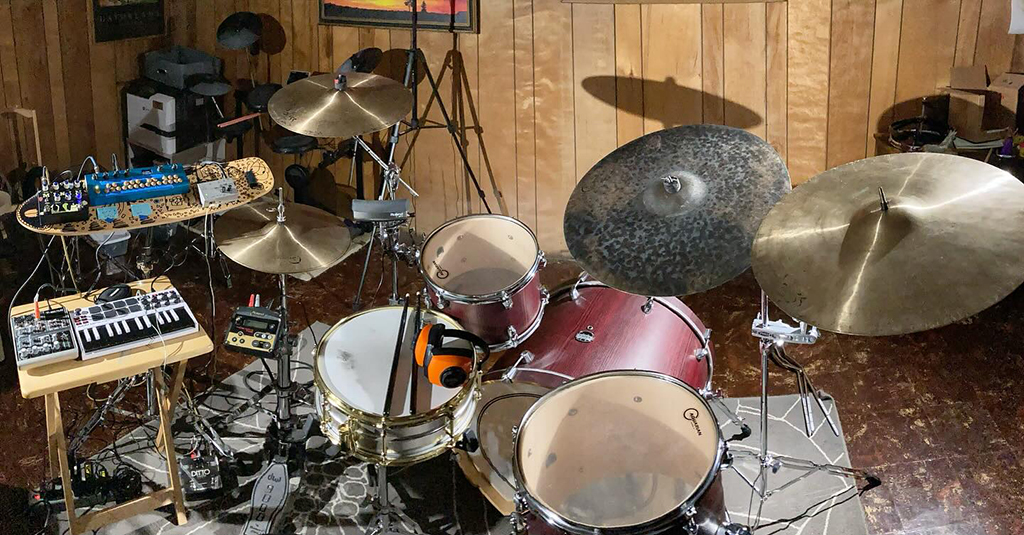 A drum set is prominently featured, with various drums, cymbals, and a pair of headphones resting on one of the drums. To the left, there’s a small keyboard on a stand and an electronic device with knobs and switches above it. The drum set includes various drums and cymbals; it’s detailed and appears professional. The setting is indoors, with wooden walls and some boxes stacked against them. The scene is lit with ambient lighting.