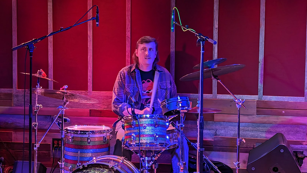 A person is seated at a drum set, illuminated by ambient lighting that casts a red hue throughout the scene. They are wearing a flannel shirt over a t-shirt adorned with a graphic design. The drum set, featuring sparkling finishes on the drums, is positioned in front of a wall with vertical wooden slats and red panels. The individual is playing a drum set that consists of various drums and cymbals. The drummer is wearing casual attire including a flannel shirt and a graphic tee. The background features wooden slats interspersed with red panels, creating an aesthetic backdrop. Microphones are positioned around the drum set for amplification. Ambient lighting casts an atmospheric glow, enhancing the visual appeal of the setting.