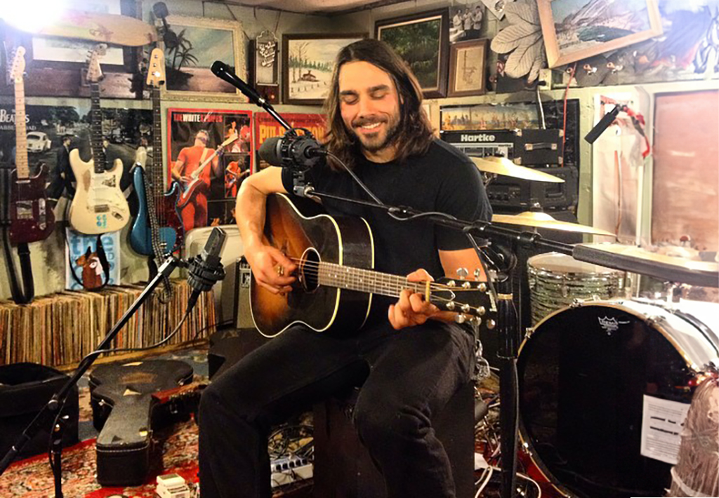 A person with long, dark hair is seated, playing an acoustic guitar. They are wearing a black t-shirt and dark pants. The individual is surrounded by musical equipment including a microphone stand in front of them and drums to their side. The setting is a room filled with various musical instruments and equipment. The walls are adorned with multiple framed pictures, guitars hanging, and other memorabilia creating a vintage aesthetic. In the background, there’s an amplifier and other electronic equipment stacked against the wall.