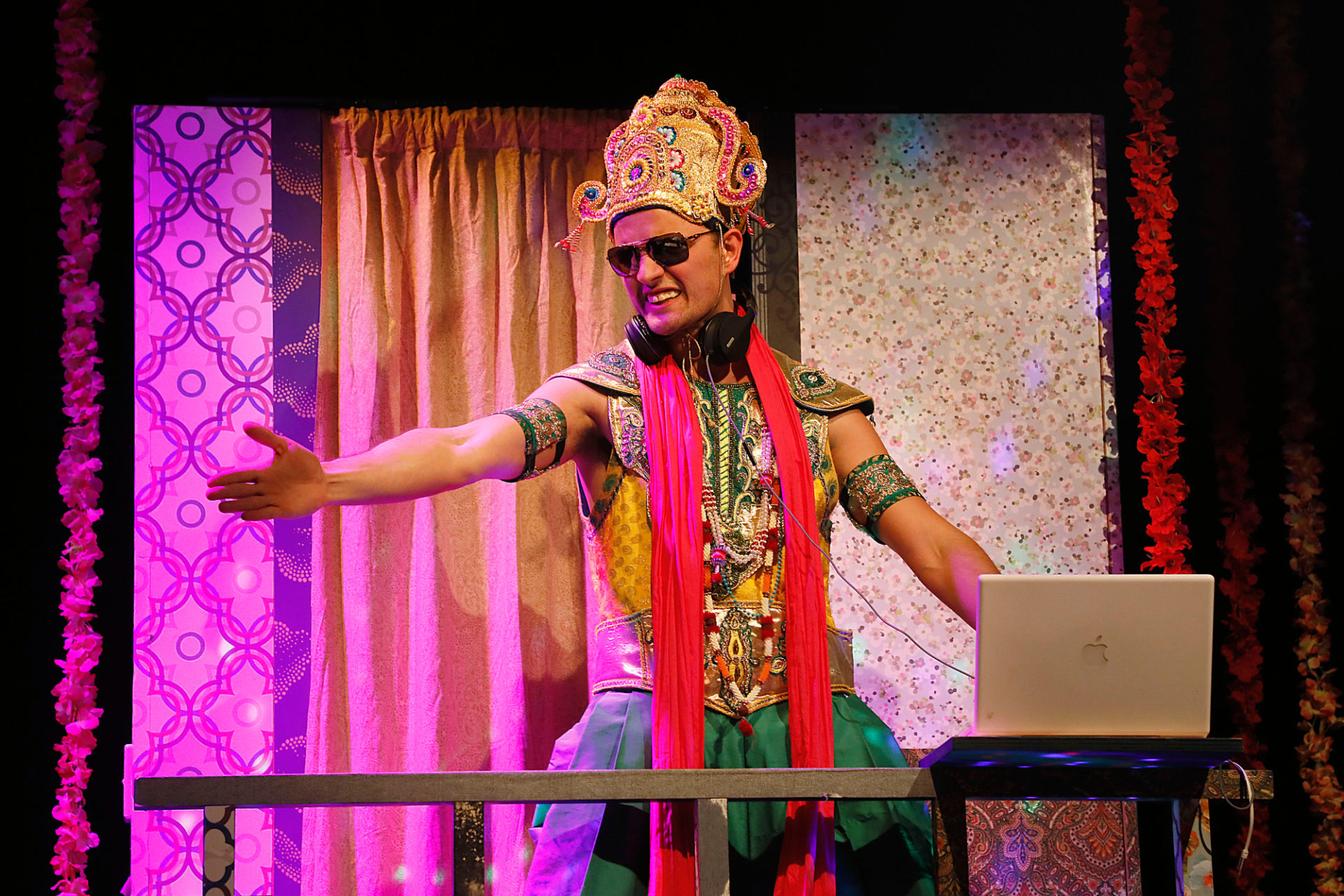 An Indian man in festival / ceremonial dress is standing behind table with a laptop on it. He is wearing sunglasses and extending one hand as if in excitement. The backdrop is colorful from the lighting design, with different fabrics serving as curtains.