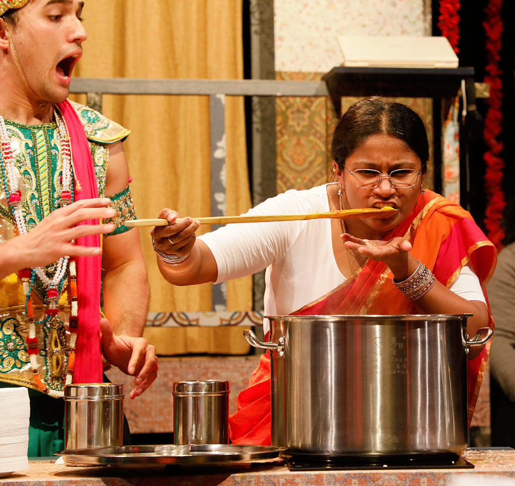 Theater production of Mrs. Krishnan's Party. An Indian man in festival/celebratory dress on the left is looking surprised with his mouth agape at an Indian woman who is leaning over a big pot and sampling food off a wooden spoon. She is wearing a white top and an orange sari.