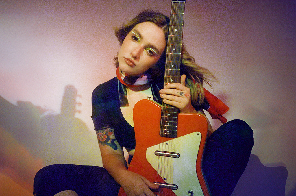 A moment is captured where a person is seated, cradling a red and white electric guitar close to their body. The individual is dressed in a black short-sleeved top and dark pants. A vibrant tattoo, full of color, decorates their left arm. Their hair, long and wavy, adds to the overall aesthetic. The background is simple, with soft lighting casting gentle shadows, contributing to an intimate atmosphere. It seems to be a quiet moment of music or perhaps preparation to play.
