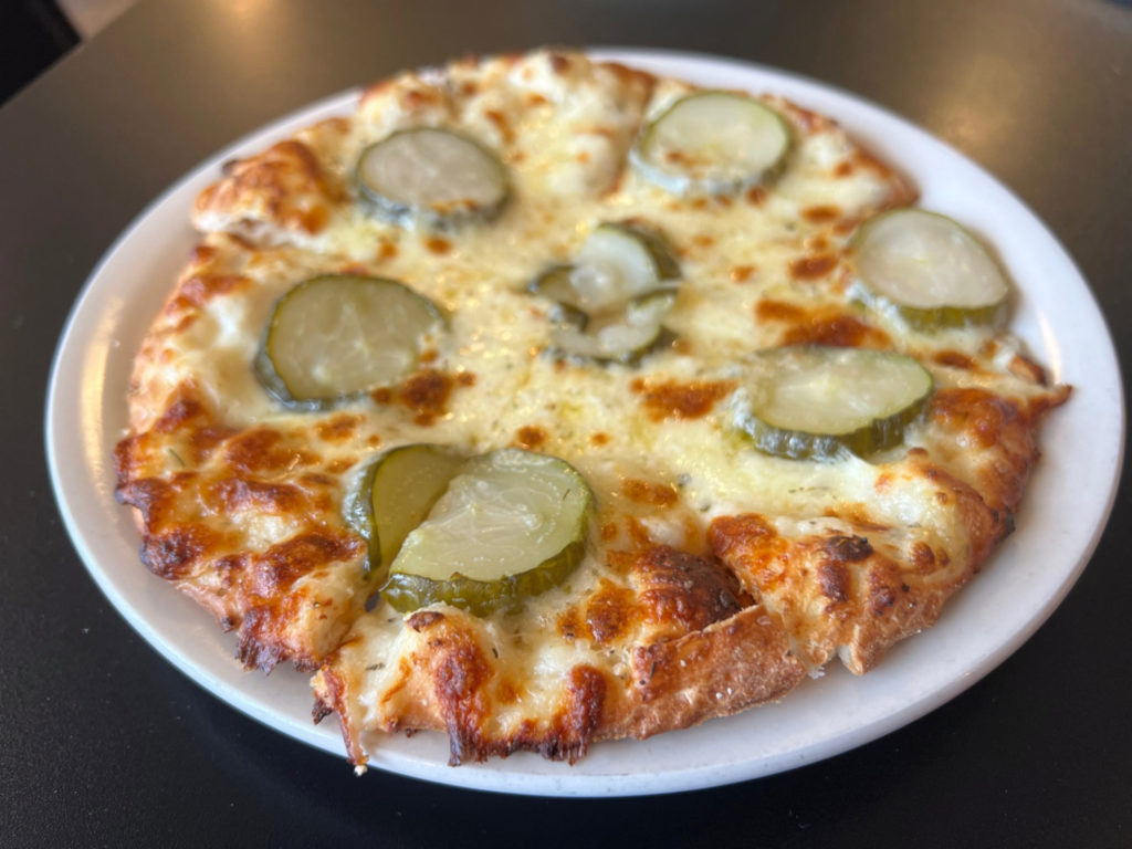 A pizza pie with garlic cream sauce and pickles, sliced but no slices taken.