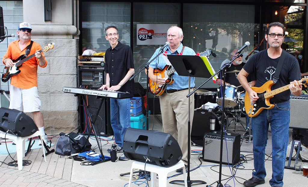 Four musicians are performing outdoors. The first musician on the left is wearing an orange polo shirt and white shorts, playing a guitar. Next to him, a person in a black shirt and dark pants is playing a keyboard. The third musician is in a light-colored long sleeve shirt and dark pants, holding an electric guitar. The last musician on the right wears a black t-shirt with white graphics and jeans, also playing a guitar. They are standing on pavement in front of a building with glass windows, with two large speakers placed on the ground in front of them. Behind them, there’s some equipment including amplifiers. The scene is set outdoors, giving a sense of a live music performance.