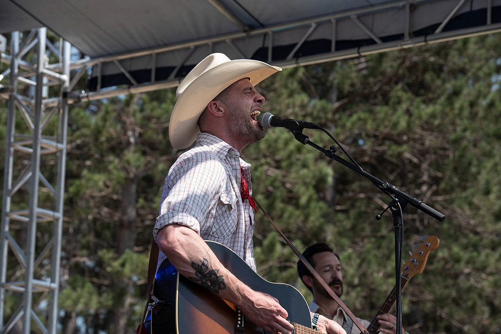 A person stands, playing an acoustic guitar. They are wearing a cowboy hat, a short-sleeve checkered shirt, and a red bandana tied around their neck. Tattoos are visible on their left arm. The setting appears to be an outdoor stage with trees in the background. Another individual is partially visible in the background, but details are not clear due to the angle of capture.