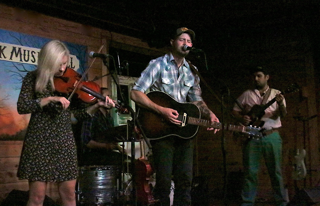 Four musicians are performing on a dimly lit stage. The person on the left is playing a violin, dressed in a dark floral dress. In the center, another musician is playing a guitar, attired in a checkered shirt and jeans. To the right, there’s another guitarist in light-colored pants and a white shirt. The backdrop of the stage has “MUSIC HALL” written on it. The lighting on the stage is dim, creating shadows around the performers.