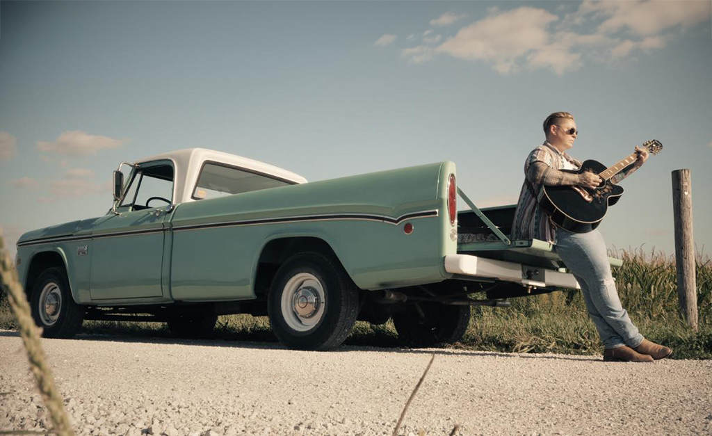 A person is standing next to a vintage pickup truck, painted in turquoise and white, parked on a gravel road. The individual is strumming a guitar and is dressed in blue jeans, a white shirt, and an open patterned button-down shirt. They are also wearing brown boots. The surrounding landscape consists of grassy fields stretching out to the horizon under an expansive sky. The scene evokes a sense of tranquility and freedom.
