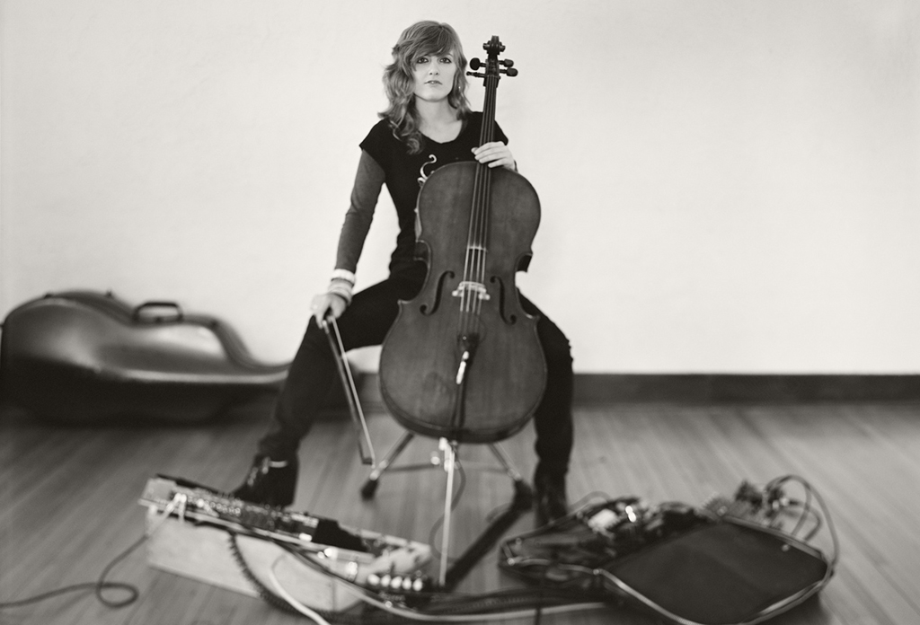 A person is seated on a wooden floor, holding a cello between their knees and grasping the bow with their right hand. They are wearing a dark, form-fitting outfit with long sleeves. The cello case lies open nearby, and an array of electronic equipment including pedals and cables is scattered around them. The setting appears to be indoors with wooden flooring and plain walls, capturing an artistic atmosphere.
