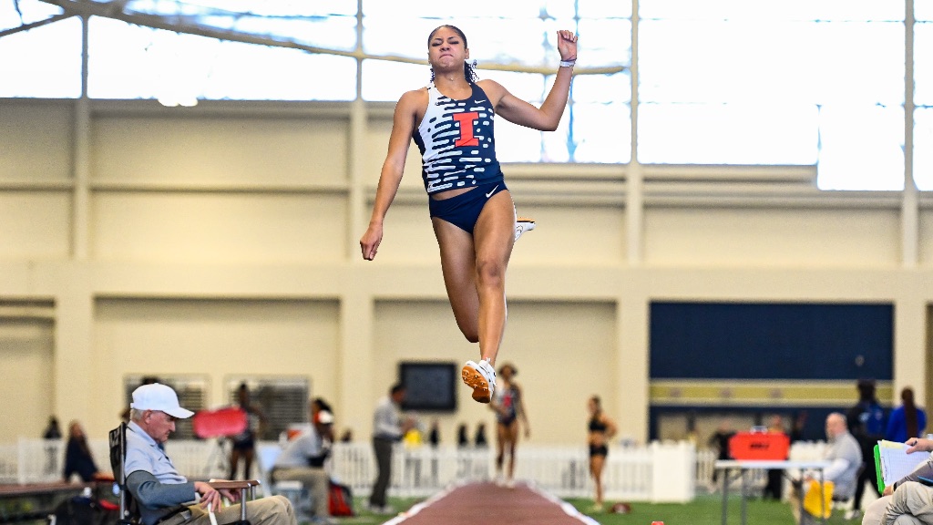 Sophia Beckman is long jumping down a long floor. She is airborne in a blue and white uniform.with an orange I.