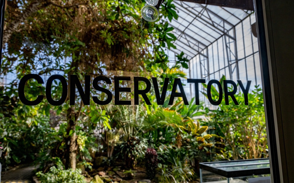 Looking through the glass door of the U of I conservatory. The word conservatory is in black upper case letters on the door. There are glass walls and the ceiling it is full of plants and trees. 