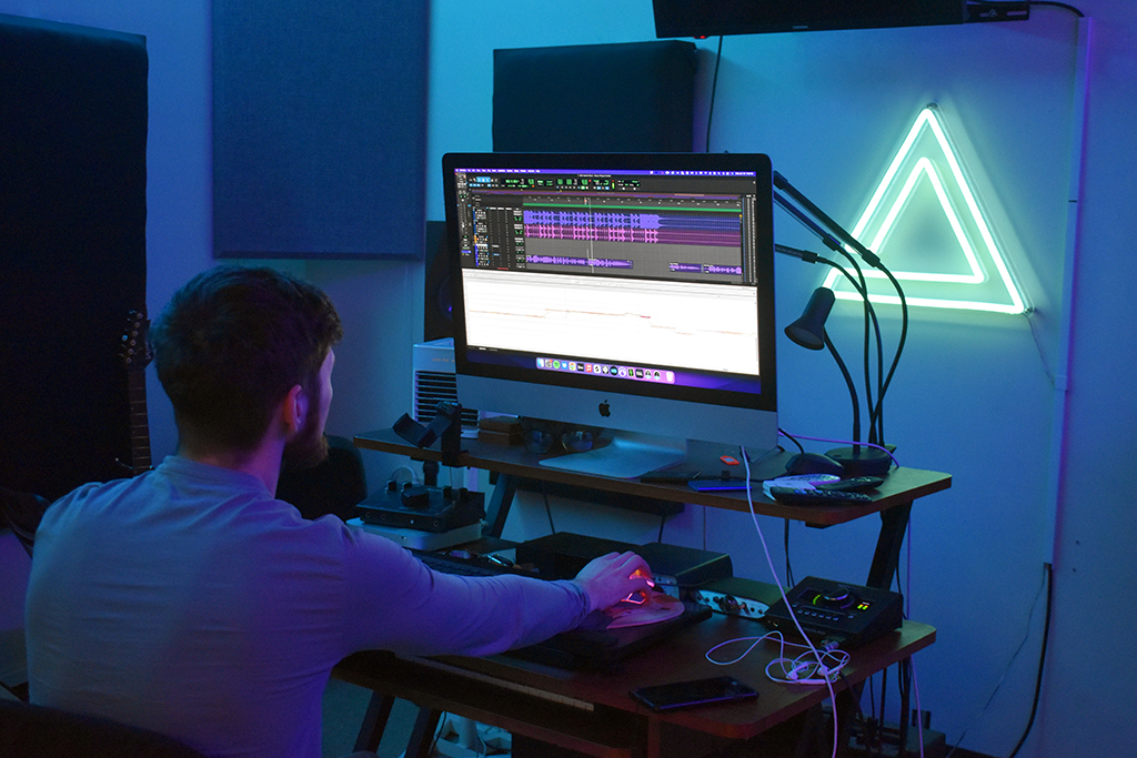 The image portrays a person seated at a desk, engrossed in work on an iMac computer. They are dressed in a long-sleeved white shirt. The screen of the computer displays a complex audio editing software interface. The room is bathed in a soft blue glow emanating from a neon triangle light mounted on the wall. This contrasts with the dark surroundings. Acoustic panels are mounted on the walls to optimize sound quality. The setting suggests a workspace equipped for audio editing.
