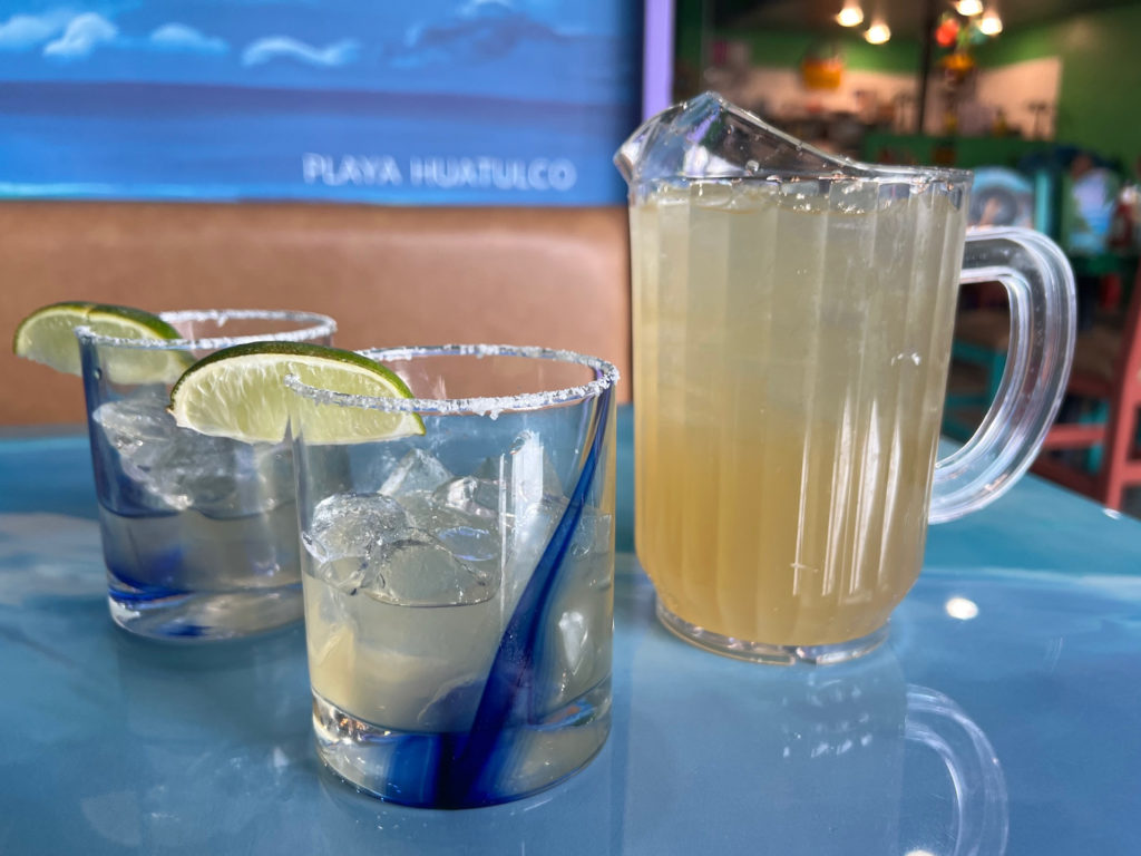 A pitcher of lime margaritas beside two half tumblers with a salted rim and lime wedge on each.