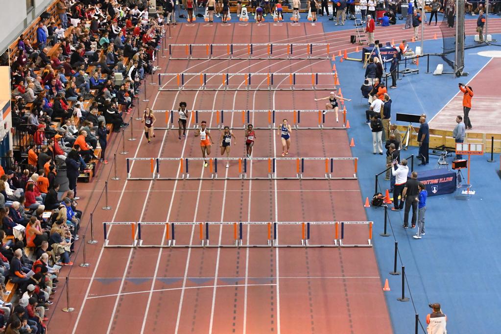 A Birds Eye view of a orange indoor track with a group of women running hurdles. There are spectators on each side of the racers.
