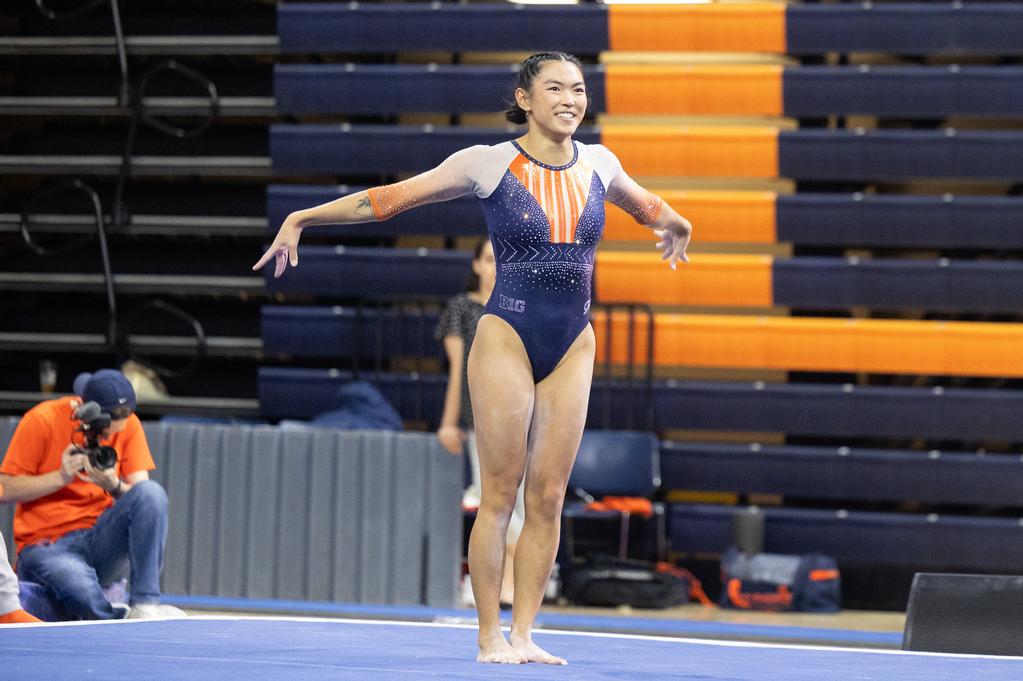 Illinois’s Mia Takekawa stands with her arms out to her sides on a blue tumbling mat during the Orange and Blue meet She has her hair in two French braids and is wearing a blue sparkly leotard with orange and white sleeves. 