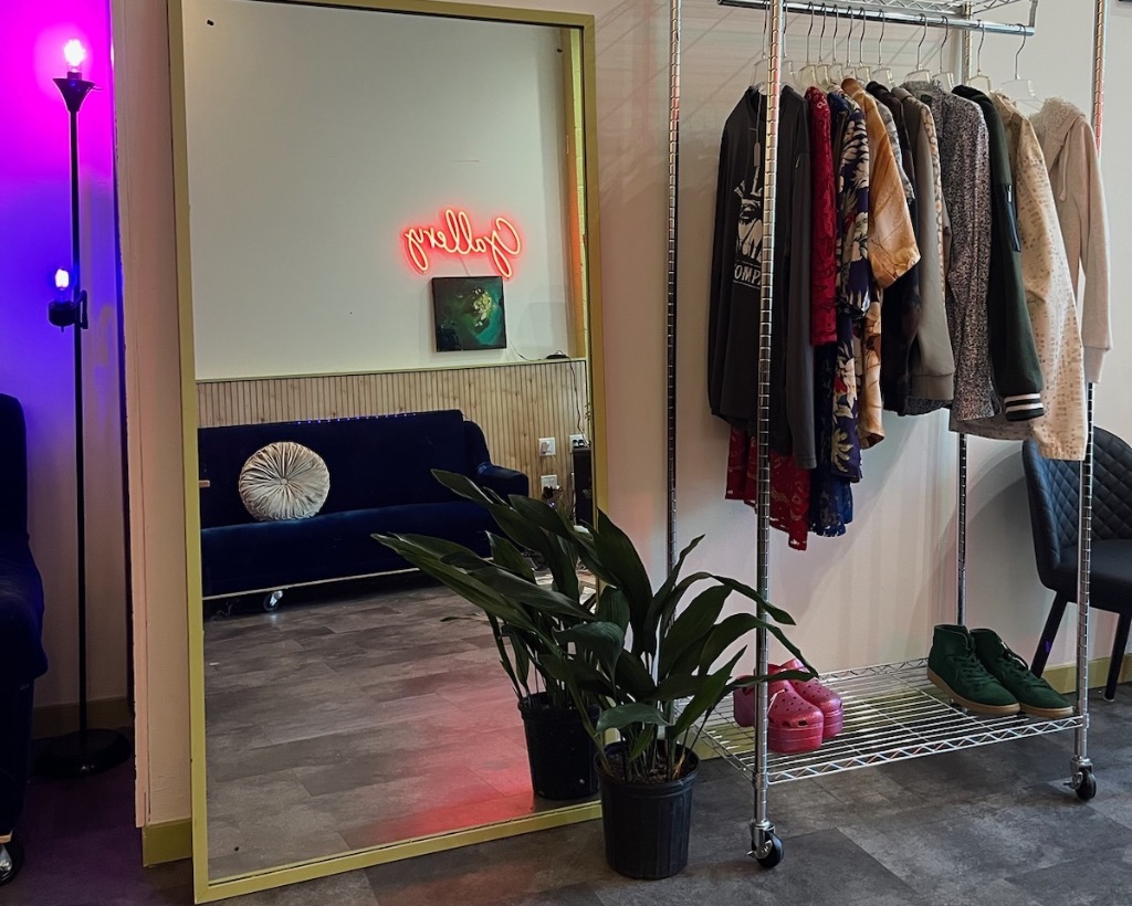 A full length mirror and rack of clothes are against a white wall. There is a plant in a gray pot on the floor and a purple and magenta light on the left side of the mirror. In the reflection art work and a neon sign are visible. 