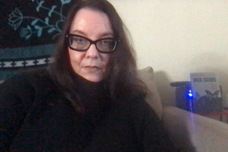 A woman with long brown hair and black-rimmed glasses, wearing a black turtleneck.