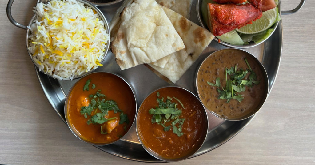 Thali lunch at Kohinoor includes chicken curry, naan, rice, lamb korma, lentils, and tandoori chicken in small metal bowls, arrangedd on a round metal tray.