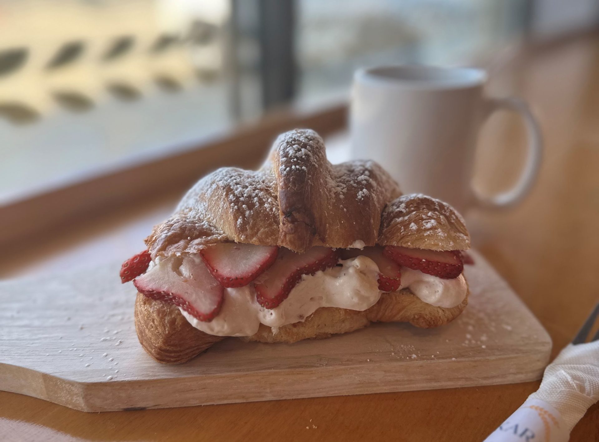 A croissant with a layer of strawberries and marscapone in the center, sitting on a wooden block. A white mug of coffee is in the background.