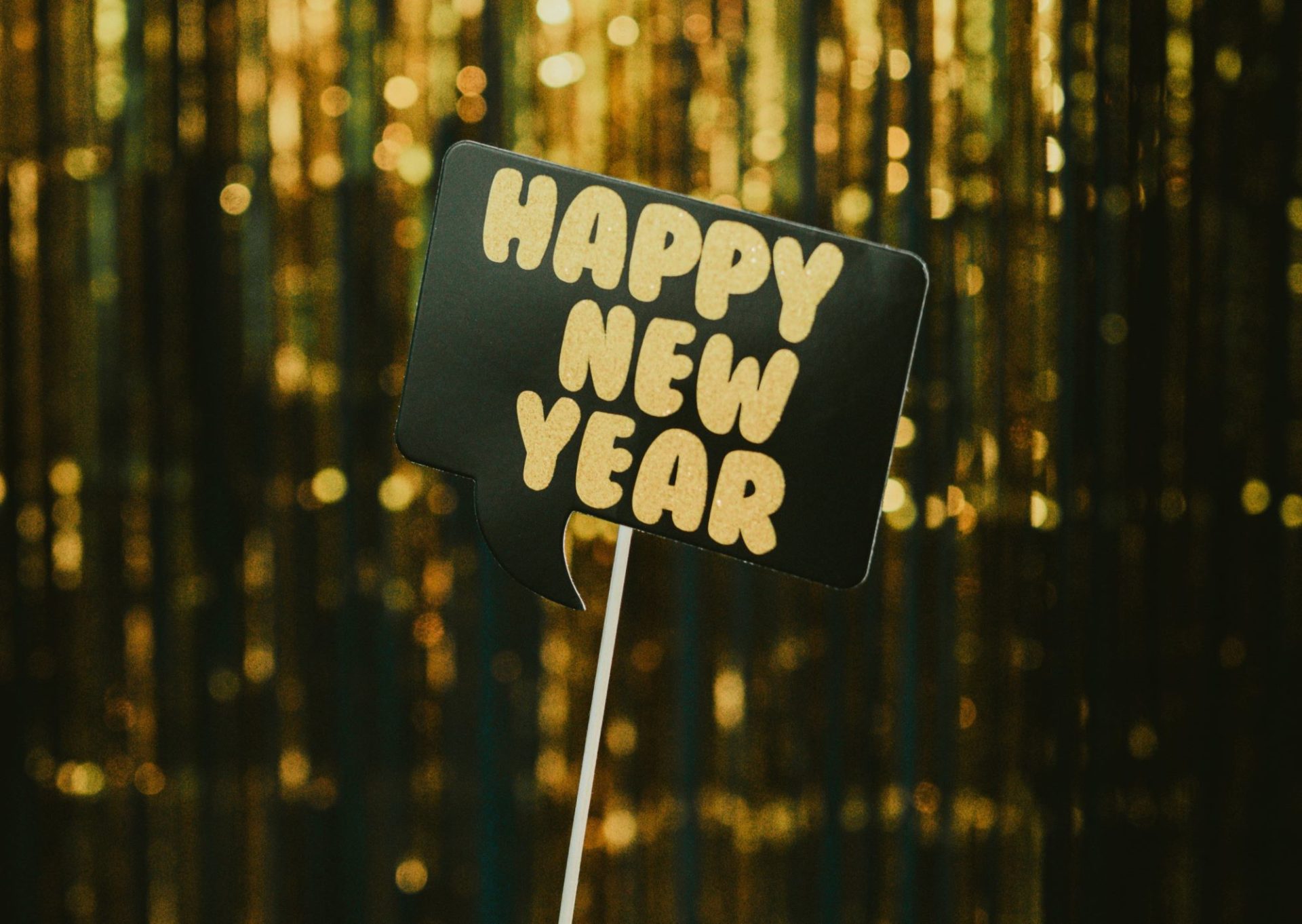 A small black sign on a wooden stick. It says Happy New Year in gold lettering. Gold tinsel hangs in the background.