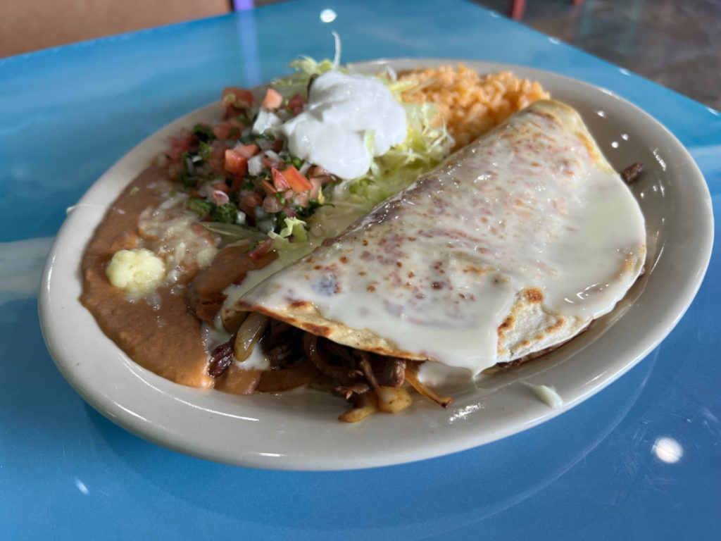 A tortilla covered in cheese and stuffed with steak is beside a side of beans, pico, sour cream, lettuce, and red rice.`