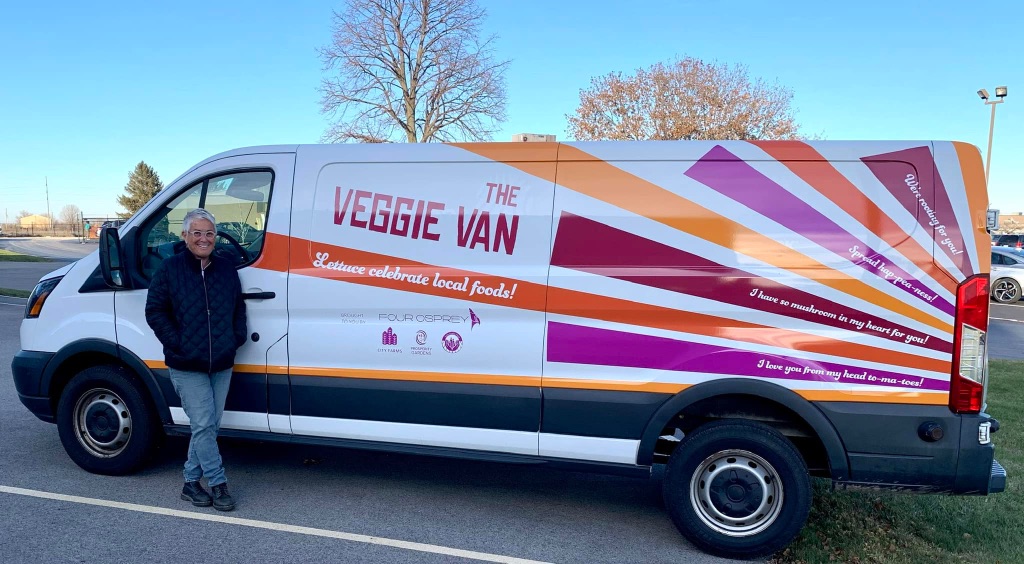A woman with short grey hair and glasses wears a black jacket and jeans and stands in front of a large white van with a sunburst of pink, orange, and maroon colors and "the veggie van" in big letters. There are a few bare trees and blue sky in the background.