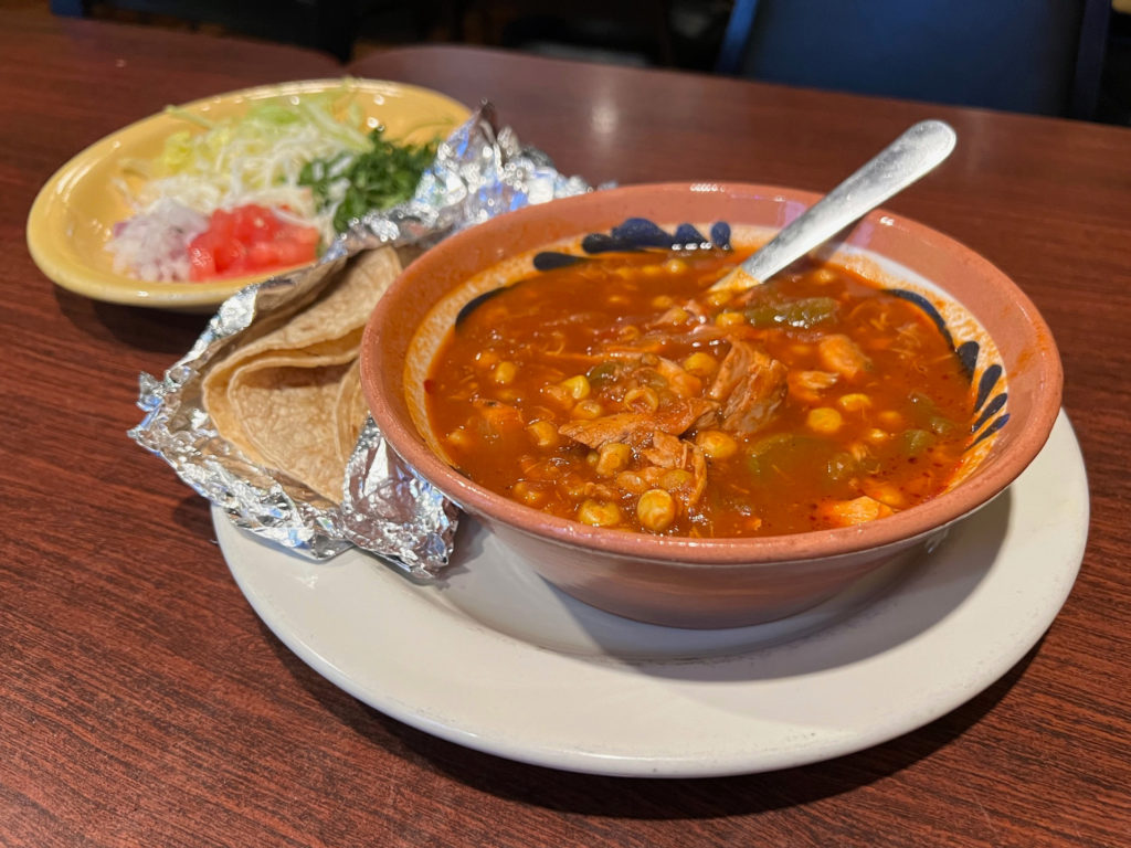 Tortilla soup in a bowl on a plate with tin-foil wrapped flour tortillas. A yellow plate holds cold condiments: red onion, tomato, cilantro, cabbage, and shredded white cheese for things to eat or drink in Champaign-Urbana.