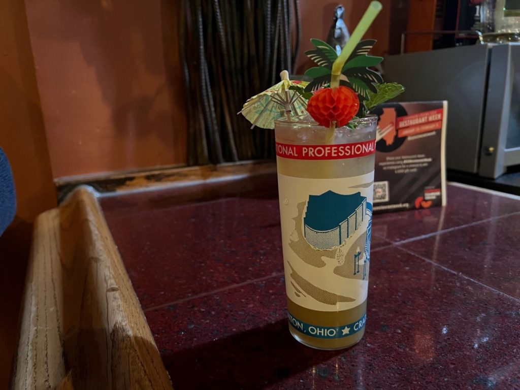 A highball glass with a yellow liquid, adorned with a yellow paper umbrella and straw with a paper pineapple. It's sitting on a brown tiled bar top.