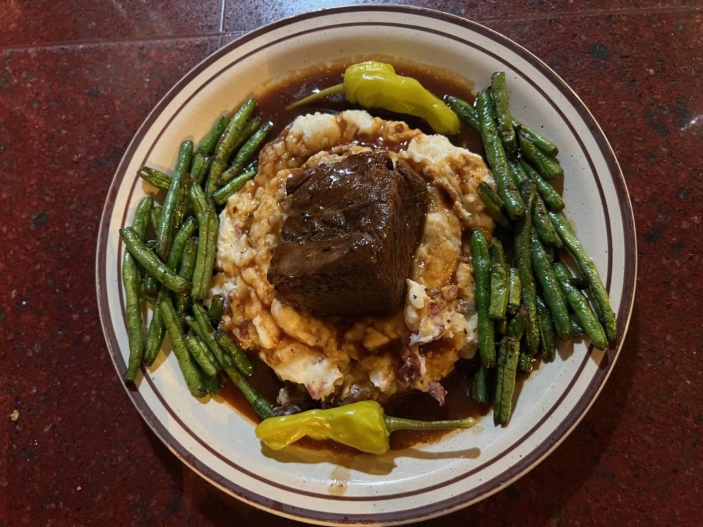 A round plate with mashed potatoes topped with a beef short rib and flanked by green beans. It's covered in a brown sauce.