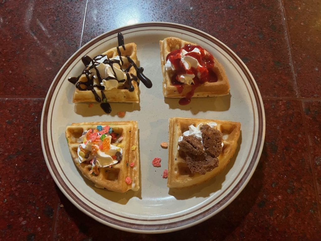 A round plate with four sections of a waffle. Each is topped with whipped cream and each has a different additional topping.