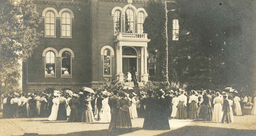 An old black and white picture of Harker Hall. There are hundreds of women in long dresses and hats with some holding umbrella. 