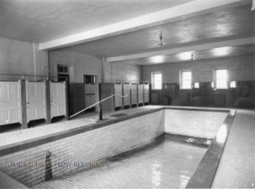 An old black and white picture of an empty swimming pool slanting downwards with changing rooms on the left side and windows on the back wall.