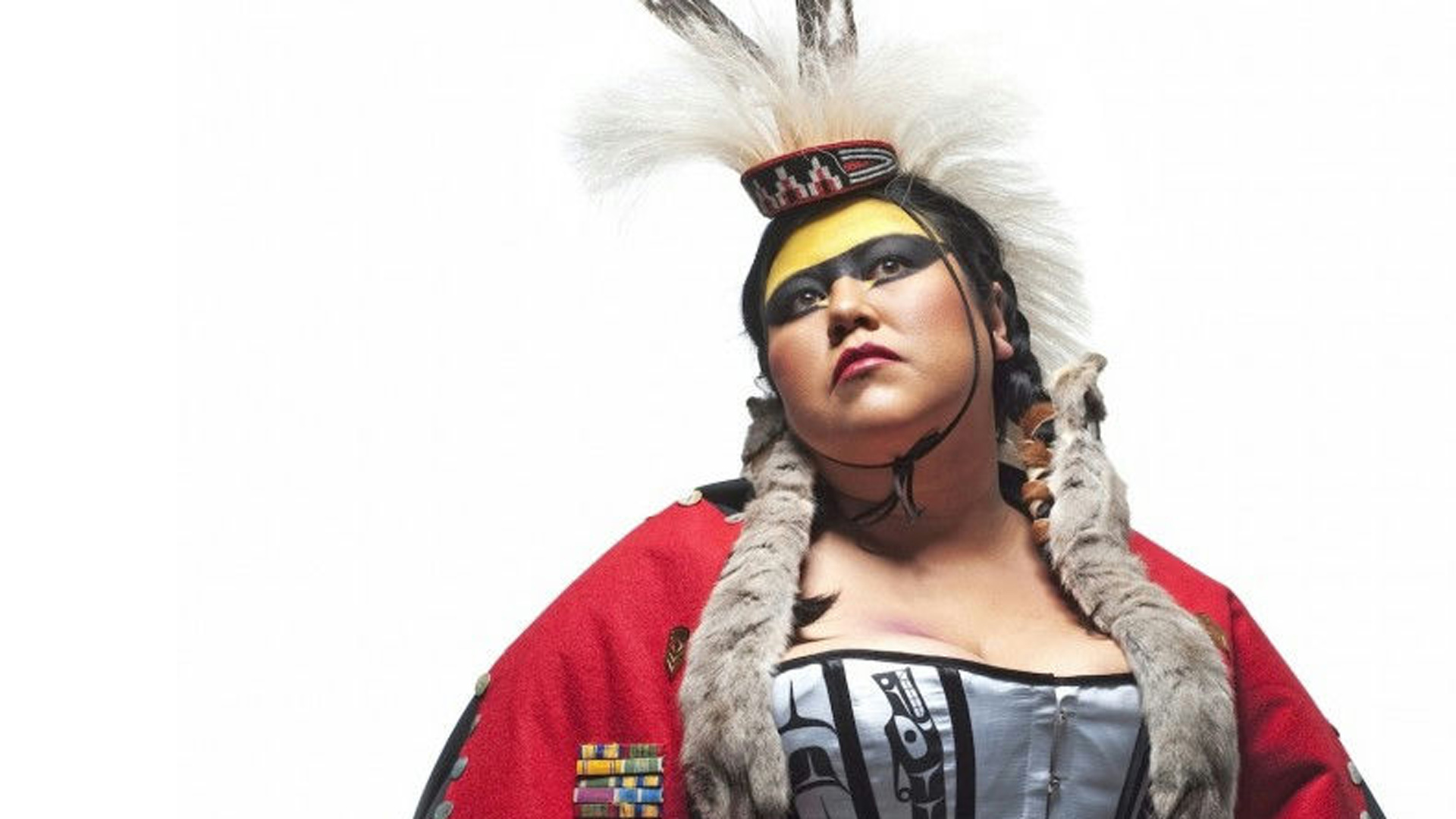 A portrait of artist Skeena Reece, a First Nations woman wearing a red robe with fur lining, and a headdress with feathers. She has yellow and black makeup across her forehead and eyes.