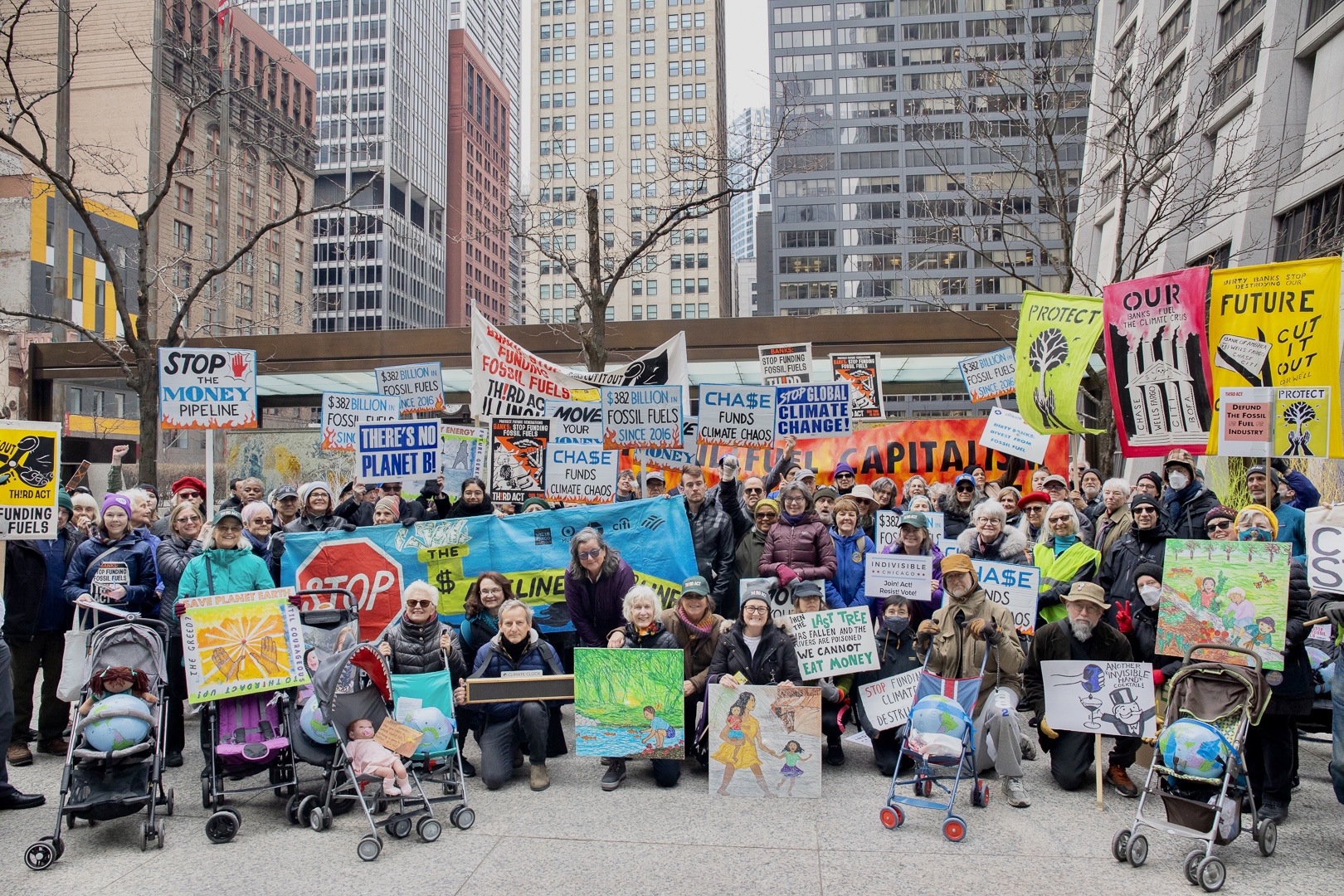 A huge group of people standing and sitting in front of the Chicago skyline. They are dressed in coats, hats, and other winter gear and holding signs encouraging the stopping of fossil fuels.