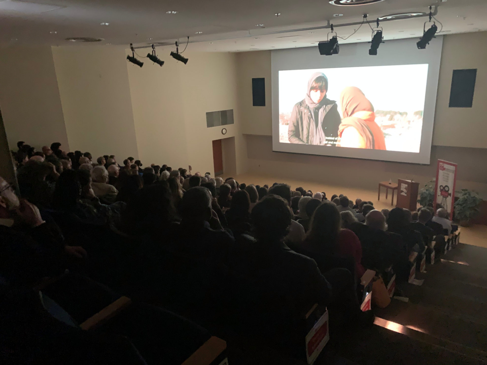 A packed audience in an auditorium watch a film screening. On the screen are two people wearing head coverings and winter coats.