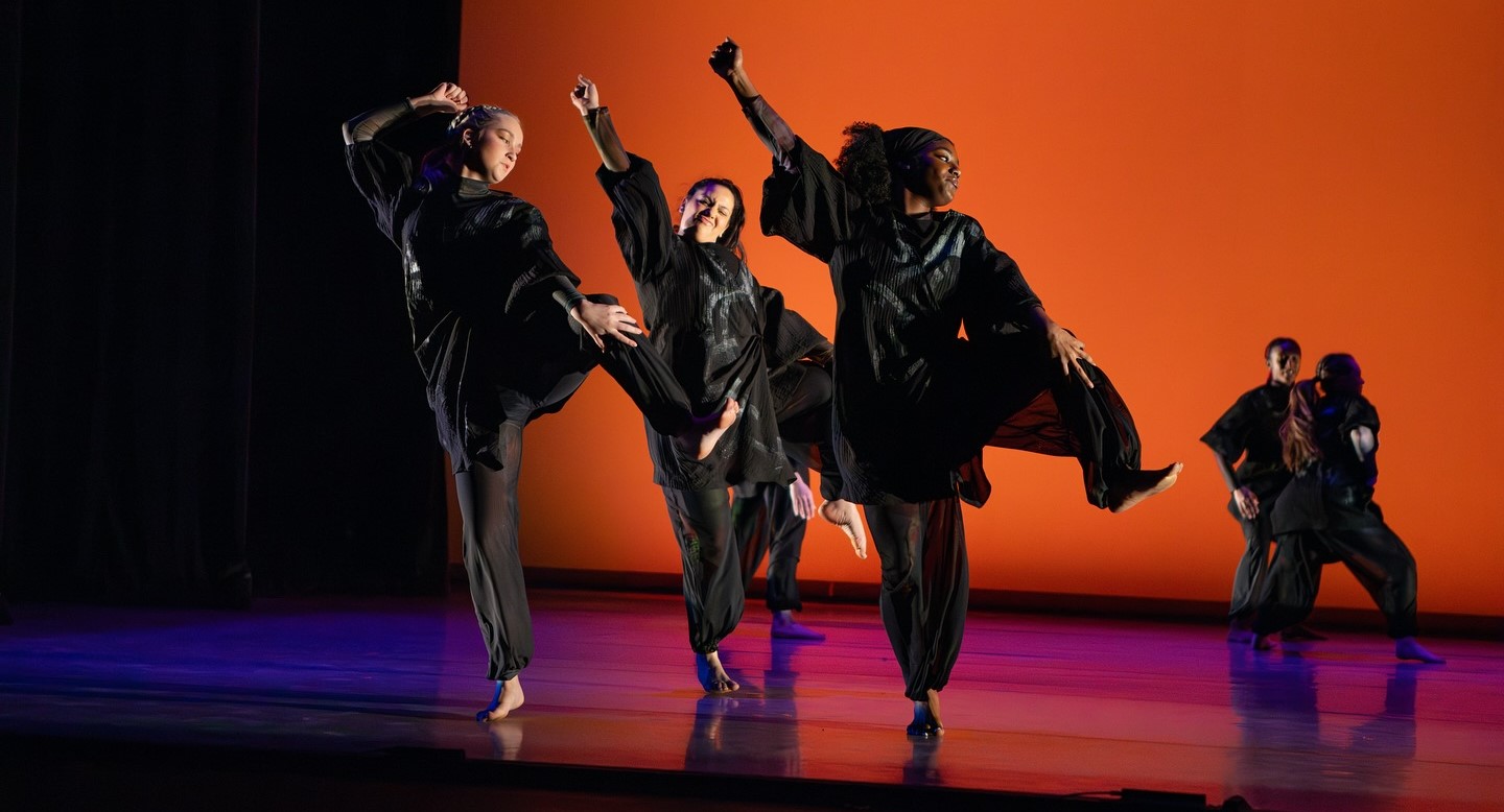 Four dancers on a stage, wearing all black and performing a dance. They each have a knee and a fist in the air.