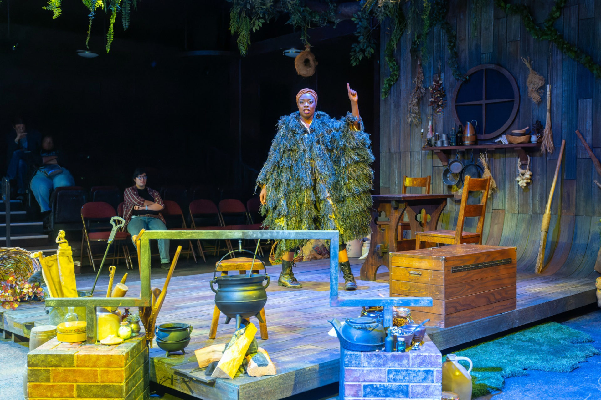 Y’vonne Rose Smith as Elizabeth in Illinois Theatre's production of Witch at Krannert Center for the Performing Arts. A black woman wears a green, fringe-covered coat and green boots. She is standing on a stage meant to resemble a 17th century house, with a cauldron over a pile of wood, a table and chairs, and a few cooking supplies.