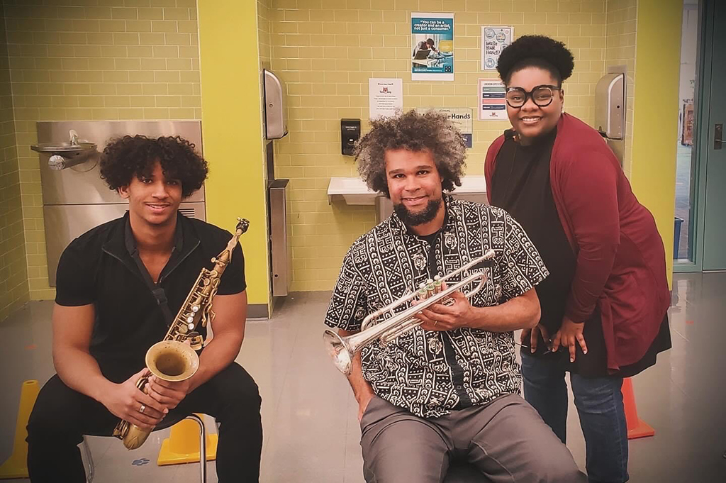 Three individuals pose for a photo in a brightly lit space with yellow-tiled walls. On the left, a person is seated on a yellow chair, holding a saxophone, wearing a black button-up shirt and black pants. In the middle, another individual is seated, holding a trumpet, and dressed in a patterned shirt and grey pants. On the right, a person stands leaning slightly towards the person in the middle, wearing glasses and a burgundy cardigan over a black top, with black pants. The background features standard fixtures of a public space, including informational posters on the wall and a hand sanitizing station.