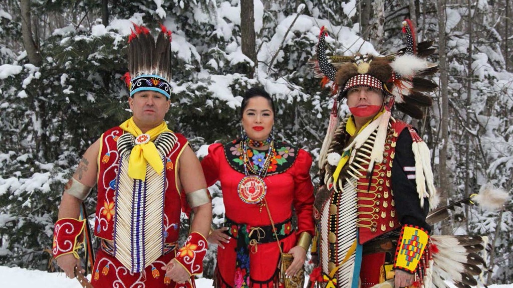 Three people stand outside against snowy pine trees. They are wearing traditional Native American clothing. It is Red, yellow, and black with beautiful beadwork, embroidery, feathers, and headdresses.