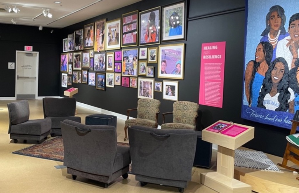 An exhibit of the Black Joy Project. A black wall is covered by many types of colorful art work In white frames. In front of the wall are grey plush armchairs. 