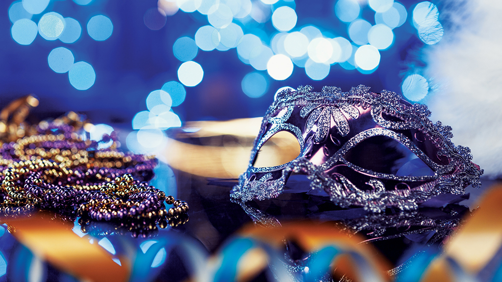 A decorative masquerade mask with intricate lace-like detailing and shimmering accents takes center stage against a backdrop of soft, bokeh light effects in hues of blue. The mask is adorned with patterns that suggest a festive and opulent design. Strands of beads in traditional Mardi Gras colors of gold and purple drape elegantly beside the mask, and a hint of feathers can be seen on the edge, adding to the festive flair. The overall composition exudes a sense of celebration and the mystery of a masquerade ball.