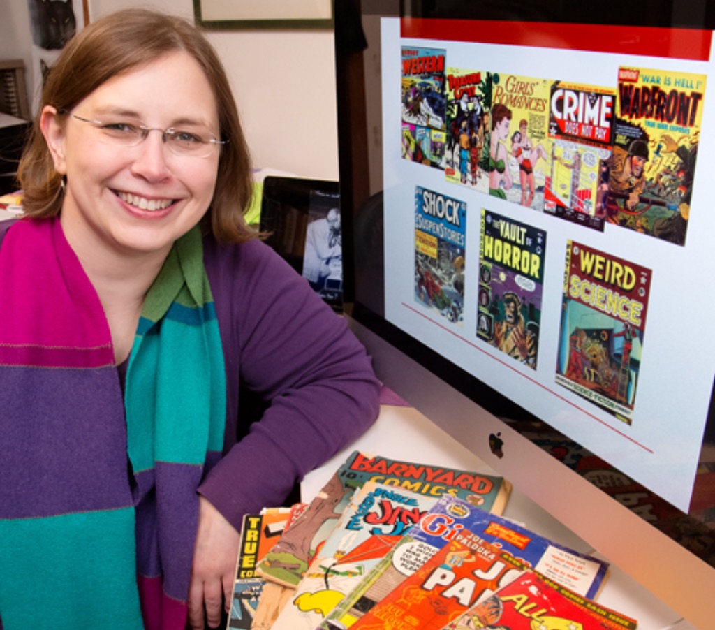 A white woman with glasses and short brown hair wears a long sleeve purple shirt wears a pink, green, and purple scarf. She sits in front of a computer with comic books on the screen. She also has comic books laid out on the desk in front of her.