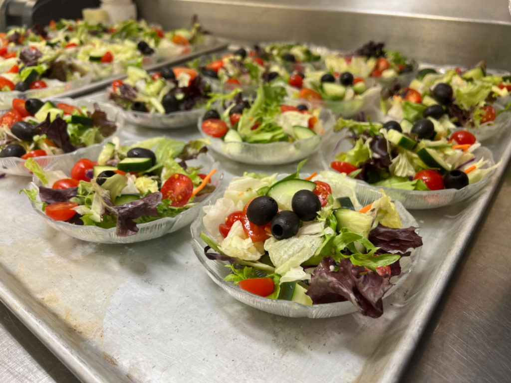 Small clear bowls of house salad with olives, cucumber, tomato, and mixed greens.